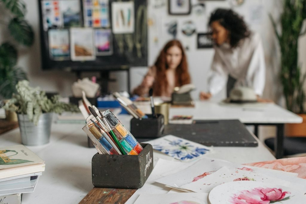 Art studio workspace with paint brushes and artists creating in the background.